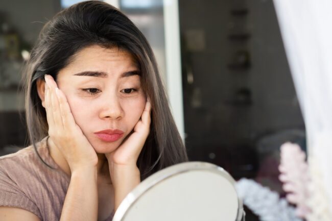 Asian woman checking her eyes bags , dark circles, wrinkle and crow’s feet on her face skin with a mirror